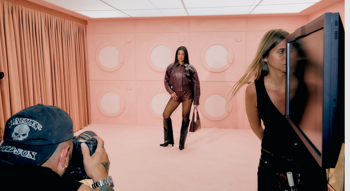 Behind-the-scenes photo of Sahar Dahi posing on a pink laundromat set in a burgundy leather jacket and shorts, black knee-high boots and a brown handbag.