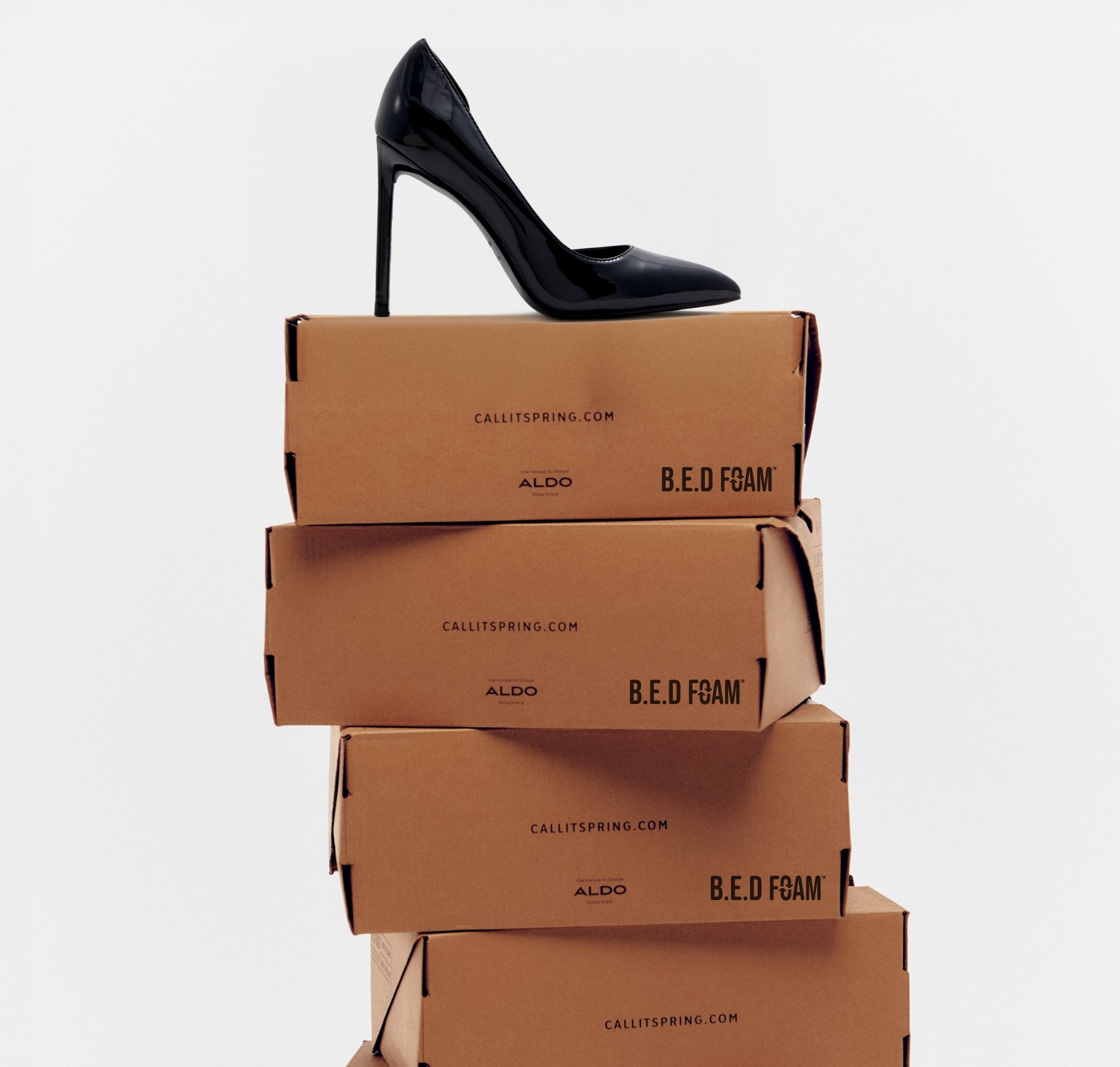 A studio product shot featuring a single black stiletto high heel shoe balanced on top of a stack of four brown cardboard shoeboxes. The boxes are printed with the 