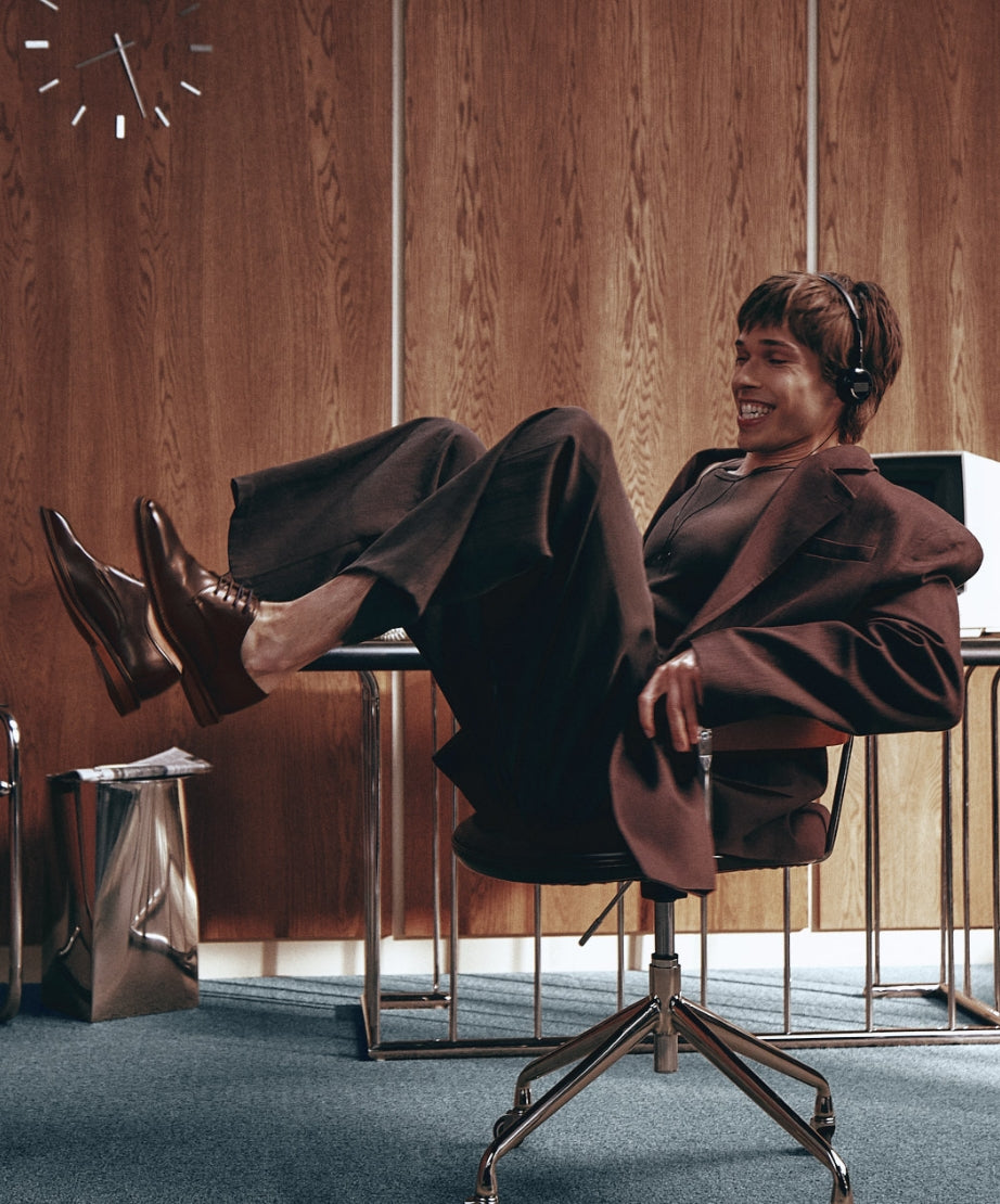 Mannequin tournant sur une chaise de bureau rétro, portant un complet brun et des richelieus bruns à lacets.