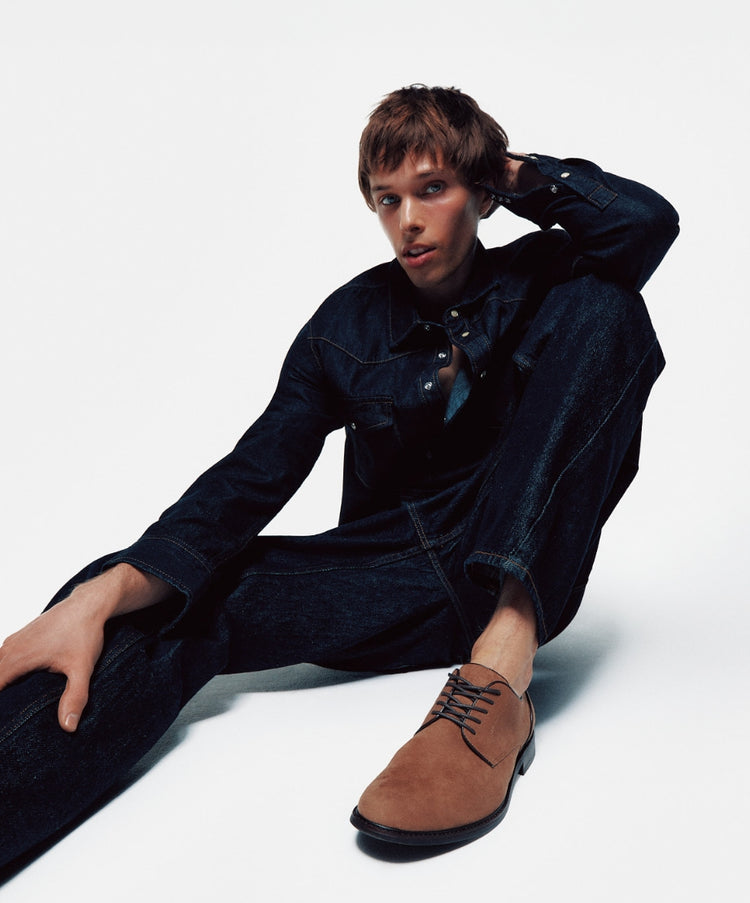 A male model wearing a denim jacket and matching jeans sitting on the floor against a light grey background, wearing brown vegan suede lace-up dress shoes.