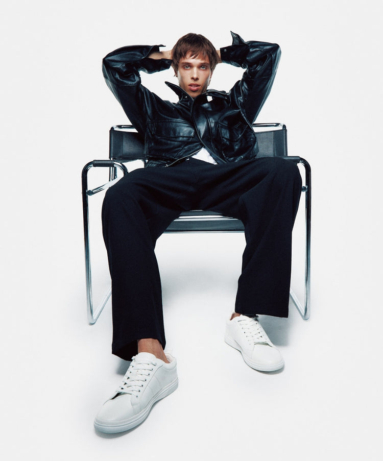 A male model wearing a dark leather jacket and pants sitting on a black metal chair against a light grey background, wearing white low-top sneakers with gum soles.