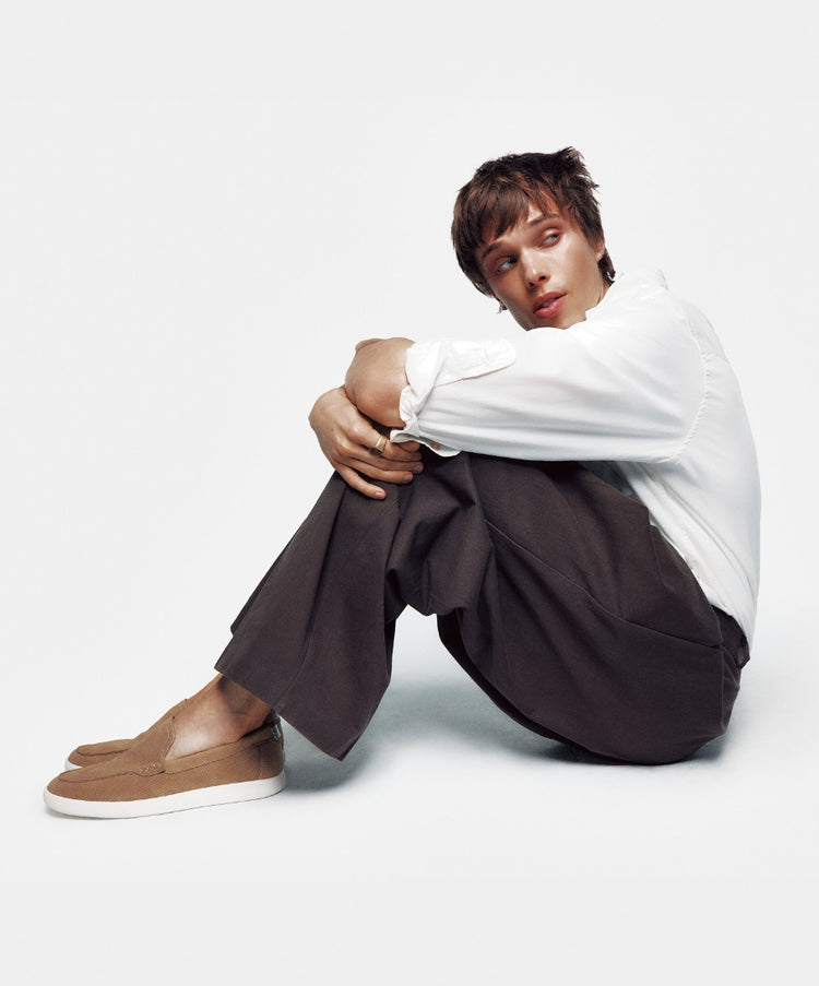 A male model wearing a white long-sleeve shirt and dark trousers sitting on the floor leaning back against a light grey background, wearing taupe suede loafers.