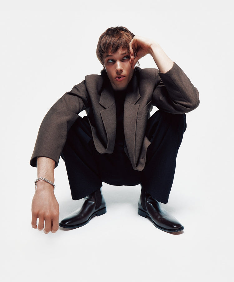 A male model wearing a dark jacket and trousers crouching against a light grey background, wearing black snip-toe vegan leather Chelsea boots.