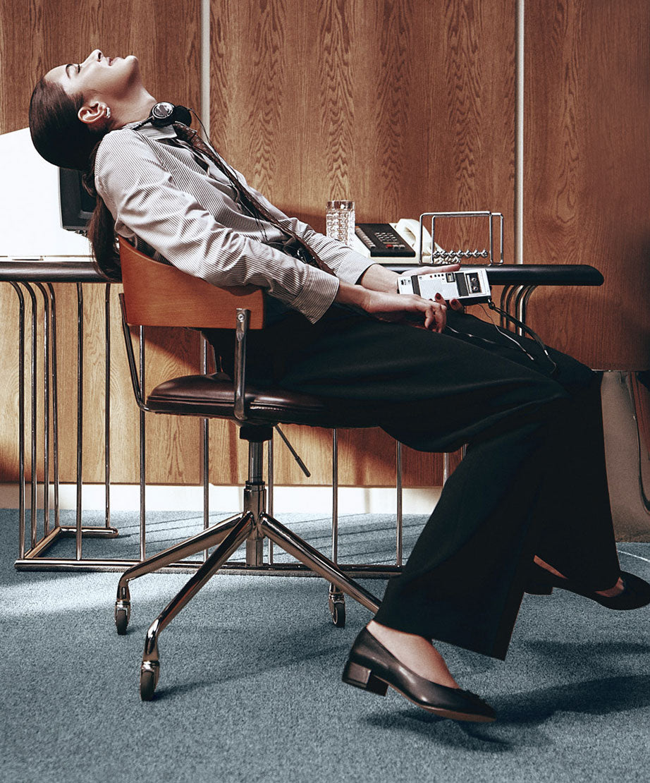 Mannequin inclinée sur une chaise de bureau rétro, portant une chemise rayée, un pantalon foncé et des chaussures plates noires.