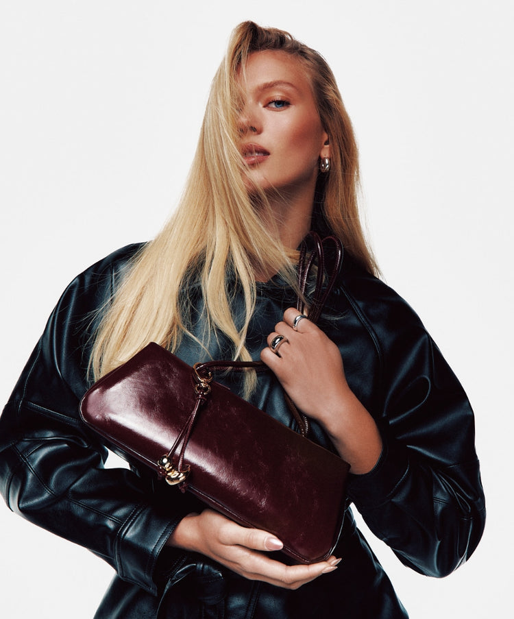 A model wearing a dark leather jacket, holding a glossy bordeaux patent leather east-west shoulder bag against a white background.