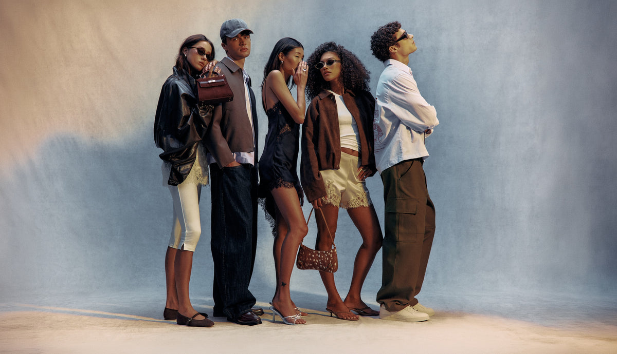 A wide group shot of five stylish young adults posing together against a textured, light grey studio backdrop.